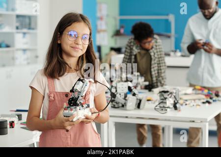 Happy schoolgirl in eyeglasses and casualwear holding handmade robot and looking at camera against her teacher and classmate Stock Photo