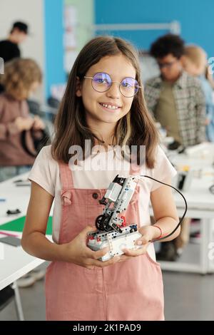 Successful schoolgirl in casualwear and eyeglasses holding robot constructed at lesson while standing in front of camera Stock Photo