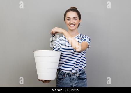 Portrait of smiling positive pretty woman wearing striped T-shirt throwing out eyeglasses to white bin, vision treatment, laser correction. Indoor studio shot isolated on gray background. Stock Photo