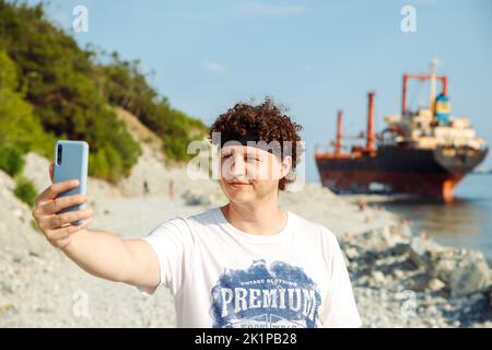 smiling Young Caucasian man with curly hair making selfie with smartphone. Guy outdoors against sunken cargo ship and taking self portrait with mobile Stock Photo