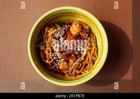 Udon stir fry noodles with vegetables, shrimps and sesame seeds in green bowl on noisy brown table mat background. Top view, close-up Stock Photo