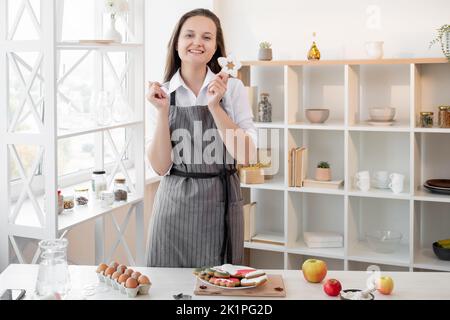 Christmas bakery. Traditional culinary. Festive cookies. Happy woman in apron showing festive decorated homemade icing biscuit light home kitchen inte Stock Photo