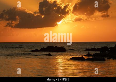 Sunset and seascape. Rocks or stones on the beach. Orange sky. Sun hidden behind clouds. Silhouette. Landscape. Stock Photo