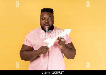 Portrait of astonished surprised man wearing pink shirt pointing direction with arrow aside, looking at camera with big eyes and open mouth. Indoor studio shot isolated on yellow background. Stock Photo