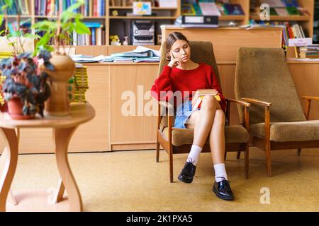 View gorgeous young woman with an interesting book in her hands. She is reading a story and sitting in an easy chair in the library in a red sweater Stock Photo