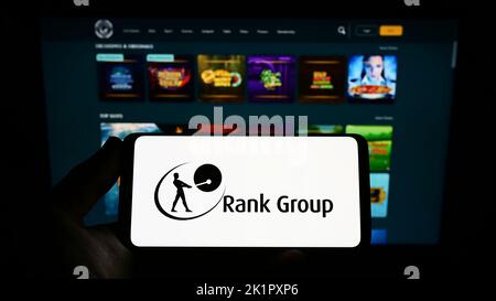 Person holding smartphone with logo of British gambling company The Rank Group plc on screen in front of website. Focus on phone display. Stock Photo