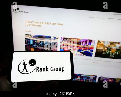 Person holding mobile phone with logo of British gambling company The Rank Group plc on screen in front of web page. Focus on phone display. Stock Photo