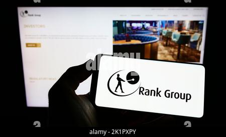 Person holding cellphone with logo of British gambling company The Rank Group plc on screen in front of business webpage. Focus on phone display. Stock Photo