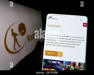 Person holding smartphone with webpage of British gambling company The Rank Group plc on screen in front of logo. Focus on center of phone display. Stock Photo