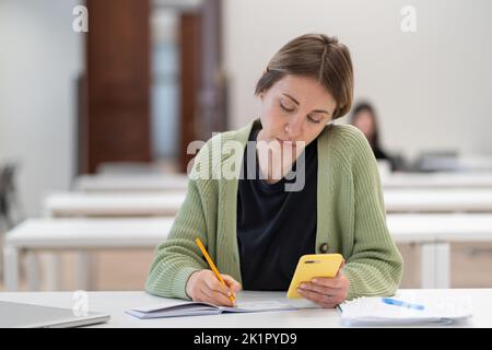 Concentrated mature female student using smartphone for exam preparation, taking second degree Stock Photo