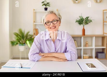 Elderly business woman with glasses smiling and looking at camera sits with hands on office table Stock Photo