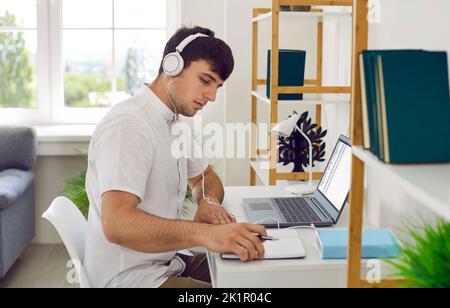 Young man working in his home office, sitting at desk, using laptop and writing in notebook Stock Photo
