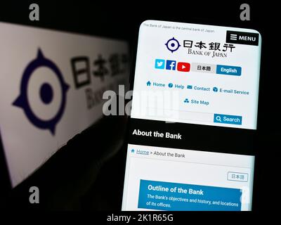 Person holding smartphone with website of financial institution Bank of Japan (BOJ) on screen in front of logo. Focus on center of phone display. Stock Photo