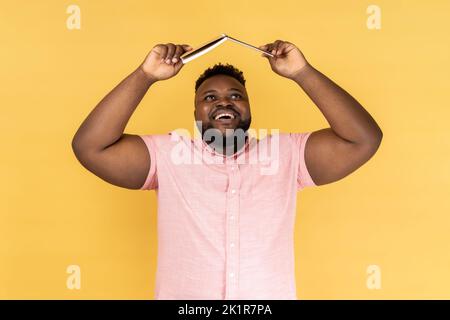 Portrait of joyful delighted bearded man wearing pink shirt holding opened book on head, looking up with happy positive expression. Indoor studio shot isolated on yellow background. Stock Photo