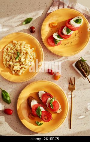 Assorted of Italian dishes with pasta fusilli, caprese salad - tomatoes, mozzarella, basil and pesto. Bread appetizer with caprese salad on orange pla Stock Photo