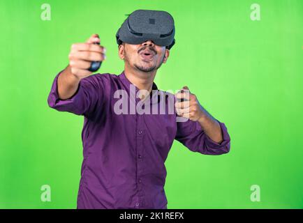 Excited young man with vr or virtual reality headset playing video game on metaverse using joystick on green screen background - concept of technology Stock Photo