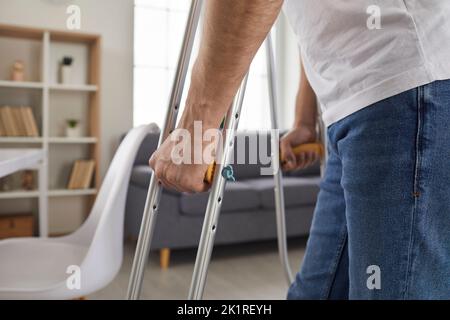 Adult man with injury walks around apartment at home, leaning on modern metal crutches. Stock Photo