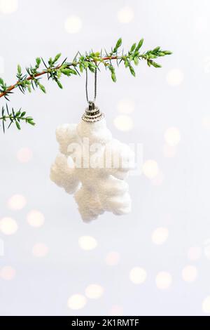 Happy holidays. White snowflake, Christmas toy hanging on green tree branch, juniper tree on white background yellow shimmering highlights. Minimal style. Vertical. Copy space. Soft Selective focus Stock Photo