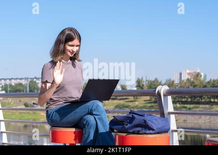 A beautiful brunette student with glasses uses a laptop on the street, chatting with friends via video chat, waving her hand in greeting. Online commu Stock Photo
