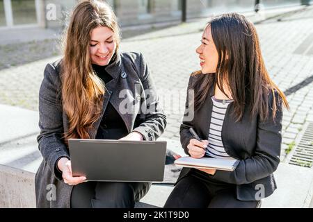 Positive woman typing on laptop while sitting near Asian female colleague writing in notepad while doing work together Stock Photo