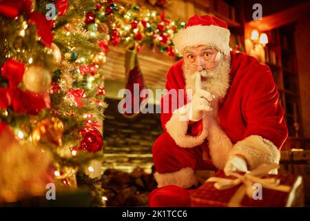 Portrait of traditional Santa Claus putting presents under Christmas tree in secret Stock Photo