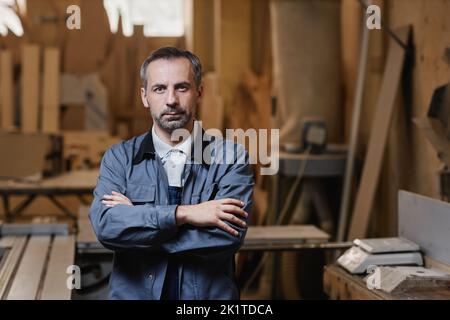 Waist up portrait of professional male worker standing in woodworking shop at factory with arms crossed and looking at camera Stock Photo