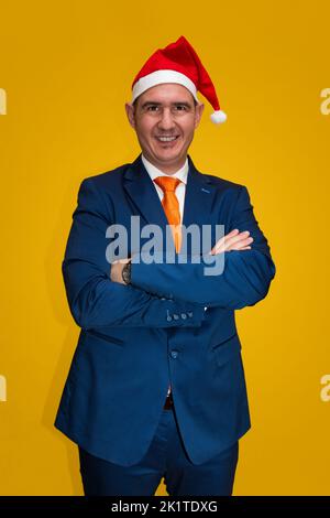 A smiling bald Caucasian adult male wearing a blue suit, white shirt and orange tie with his arms crossed is wearing a red and white Santa Claus hat o Stock Photo