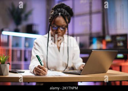 Cheerful confident african american business woman, office manager, wearing white shirt using laptop while making financial report while writing on paper working at night. Stock Photo