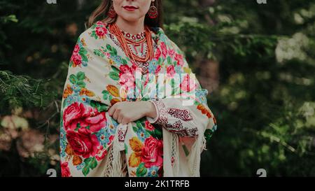 Pretty woman in traditional ukrainian necklace, dress, handkerchief on green nature backdrop. Lady in national costume - vyshyvanka and shawl with Stock Photo