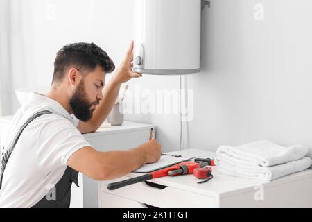 Male plumber writing on clipboard near boiler in bathroom Stock Photo