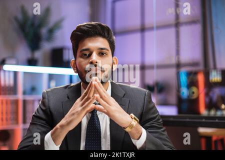 Indian bearded man in formal suit sitting at desk with modern laptop and looking at camera with serious facial expression. Concept of people and office work. Stock Photo