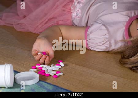 Unconscious little child (girl) with pink pills laying on the floor at home. Danger of medicament intoxication. Stock Photo