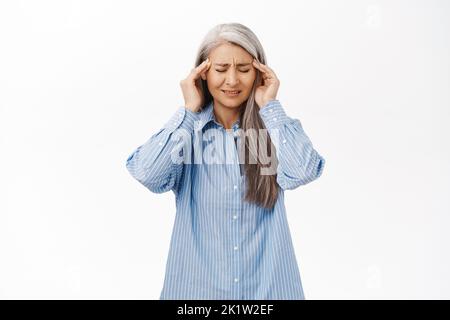 Portrait of japanese senior woman having headache, touching head and grimacing from pain, migraine, standing over white background Stock Photo