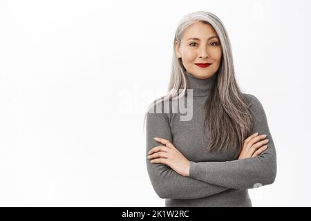 Portrait of beautiful, healthy smiling middle aged asian woman, cross arms on chest, looking confident and happy, standing over white background Stock Photo