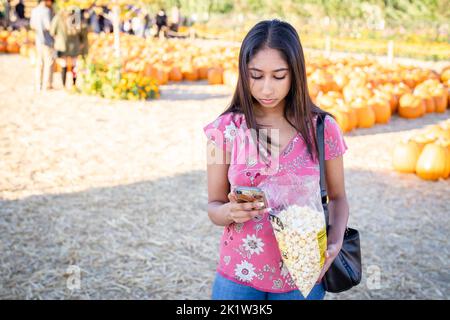 Fall Celebration Portrait of a Young Woman Eating Kettle Corn at a Pumpkin Farm while on Her Smart Phone Stock Photo