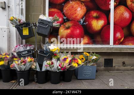 Bunches of fresh cut flowers in black plastic tubs outside a fruit and vegetable shop with red apples poster. Stock Photo