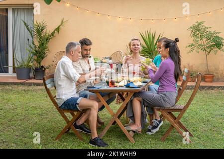 Mature friends eating and drinking happy at dinner barbecue outside in the backyard. Middle-aged people during weekends. Stock Photo