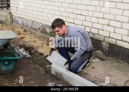 Defocus young man laying gray concrete paving slabs in house courtyard on gravel foundation base. Master lays paving stones. Repairing sidewalk. Out o Stock Photo