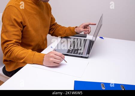 A shot of a man sitting in front of a laptop while holding a pen to write something on a piece of paper Stock Photo