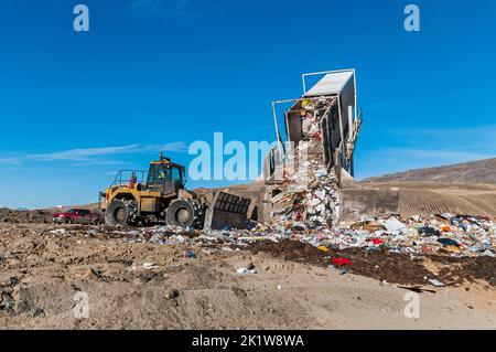 A trailer from a semitrailer truck is in a landfill tipper being raised and dumps out trash, while a soil compactor bulldozer pushes the trash. Stock Photo