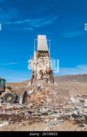 A trailer from a semitrailer truck is in a landfill tipper being raised and dumps out trash, while a soil compactor bulldozer pushes the trash. Stock Photo