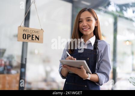 Charming Asian businesswoman in an apron hold an order book in front of a cafe counter with a beaming smile preparing to open and take orders. Stock Photo