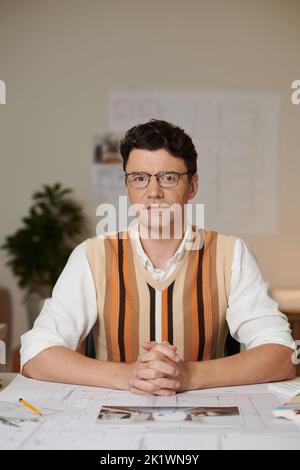 Portrait of serious confident architect in glasses sitting at desk with blueprints and looking at camera Stock Photo