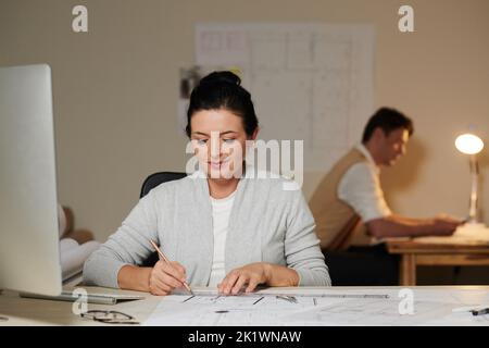 Smiling mature female engineer checking building blueprint on table Stock Photo