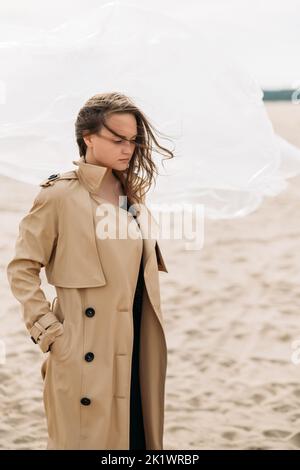 Fashion portrait. Female tenderness. Autumn melancholy. Pensive woman in beige trench coat with flying away hair in cold wind standing alone on sandy Stock Photo