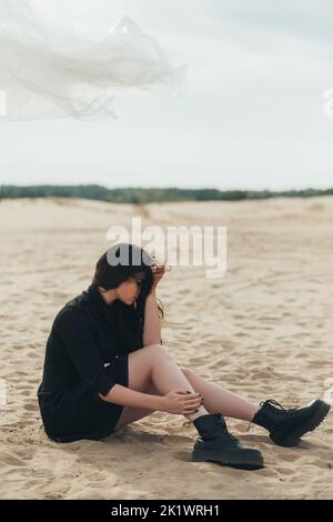 Desperate woman. Female loneliness. Emotional crisis. Sorrow grief. Depressed upset brunette lady in black casual dress sitting on sand in deserted be Stock Photo