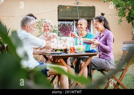 Group of mature adult friends talking and having fun in a dinner party at backyard, Men and women eating healthy food and drinking wine. Lifestyle Stock Photo
