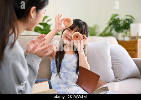 A cute and playful young Asian girl holding doughnuts, playing peekaboo with her mom while relaxing in the living room together. Happy family moments Stock Photo