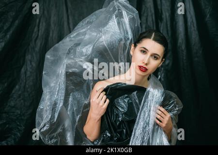 Fashion portrait. Aesthetic cosmetology. Plastic surgery. Female model with perfect face skin night makeup red lips posing in wrinkled polyethylene fi Stock Photo