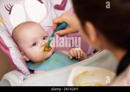 Mom feeds her baby girl with a spoon on baby chair. Stock Photo